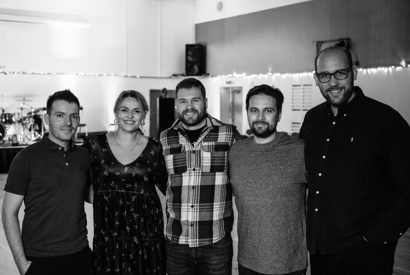 Black and white photo of five people standing close together indoors, smiling at the camera. They are in a casual setting with string lights in the background and music equipment visible on the left.